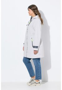 Veste imperméable gris clair avec capuche, rayures navy aux poignets et poches latérales zippées. Portée sur un jean bleu et des baskets grises.