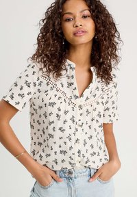 Short-sleeve button-up shirt in cream, featuring a floral pattern in black, with lace detailing across the chest and a relaxed fit.