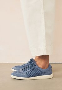 Sneakers in suede blu con una texture intrecciata sui lati, suola in gomma bianca e allacciatura standard. Abbinati a pantaloni bianchi alla caviglia.