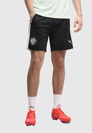 Homme portant un short de football noir St. Pauli, des chaussettes blanches Puma et des crampons Puma orange vif, debout avec une main dans la poche.