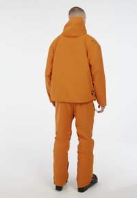 Narančasta skijaška jakna i hlače opuštenog kroja. Jakna ima podesivu kapu; hlače imaju zatvarače na gležnjevima radi lakšeg oblačenja.