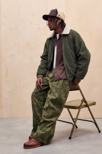 Giubbotto bomber in corduroy verde, camicia a righe marroni, pantaloni verdi a fantasia e mocassini marroni. Seduto su una sedia pieghevole contro uno sfondo semplice.