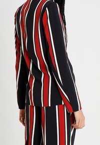 Blazer à rayures noir, rouge et blanc. Présente des rayures verticales, une coupe ajustée, des manches longues et une texture de tissu lisse.