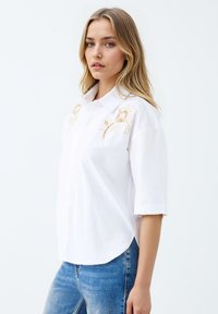 Weiße Bluse mit floralem Stickerei in Beige an den Schultern, kurzen umschlagenen Ärmeln und einem abgerundeten Saum. Getragen mit verwaschenen blauen Jeans.