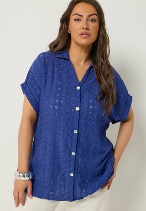 Femme portant une chemise bleue à manches courtes avec boutons œillets blancs et un gros bracelet argenté, posant avec la main sur la hanche.