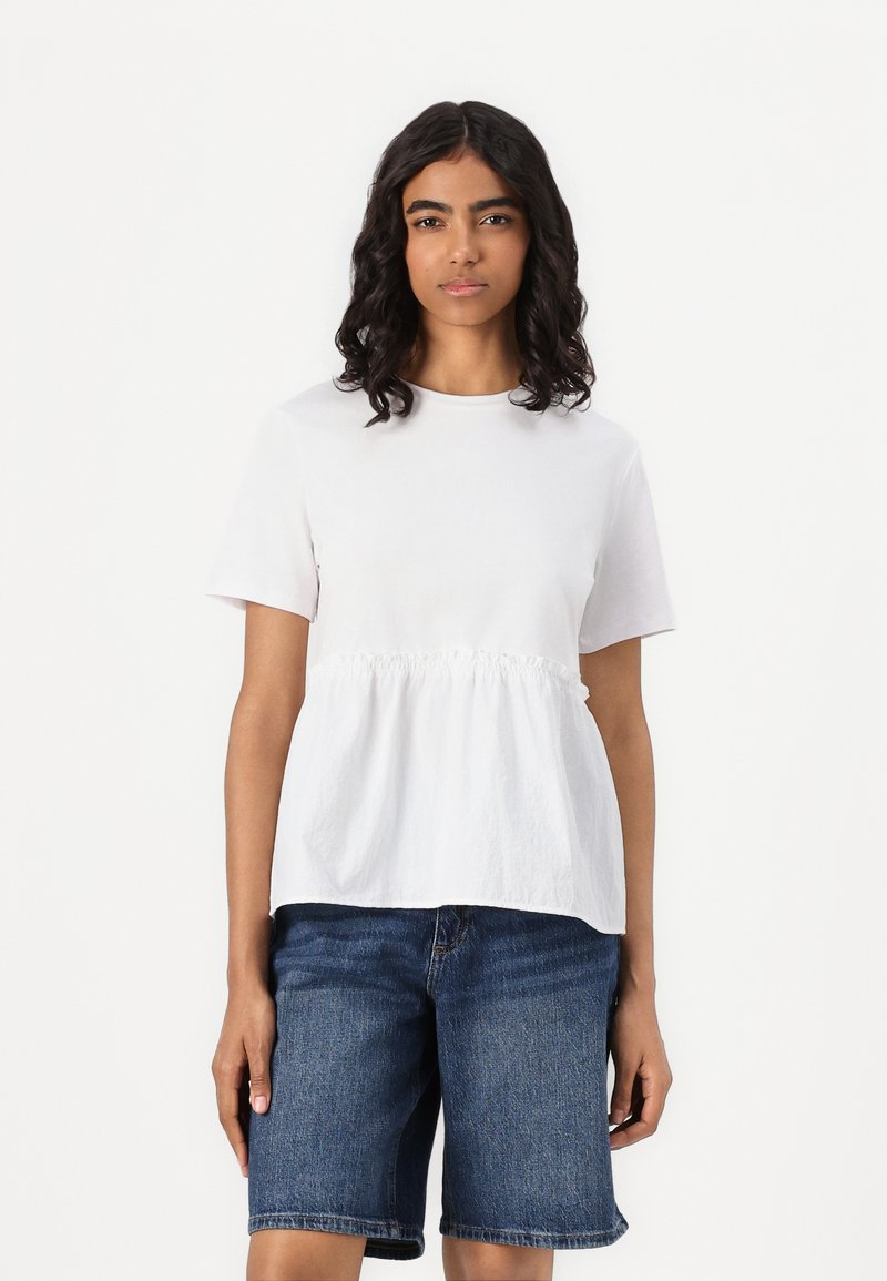 Hvit kortermet topp med detaljerte samlede midje, kombinert med blå denimshorts. Tøyet ser lett og teksturert ut.