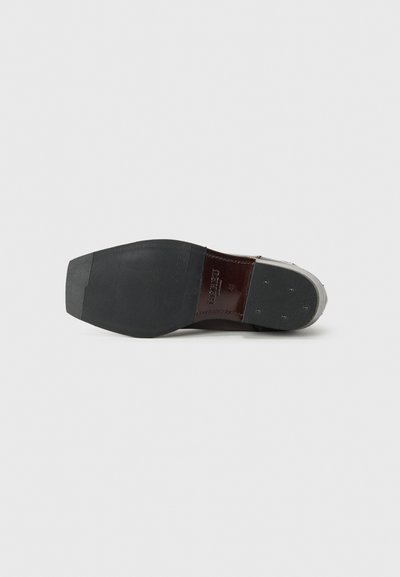 Suela de zapato de cuero con un patrón texturizado de goma negra, que presenta una zona del talón de cuero marrón liso y pequeños clavos metálicos para mayor tracción.