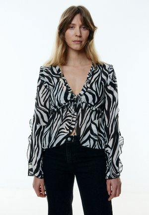Durchscheinende Bluse im Schwarz-Weiß-Zebra-Druck mit gerafften Kanten, V-Ausschnitt und versammeltem Bindedetail an der Vorderseite, kombiniert mit schwarzen Jeans.