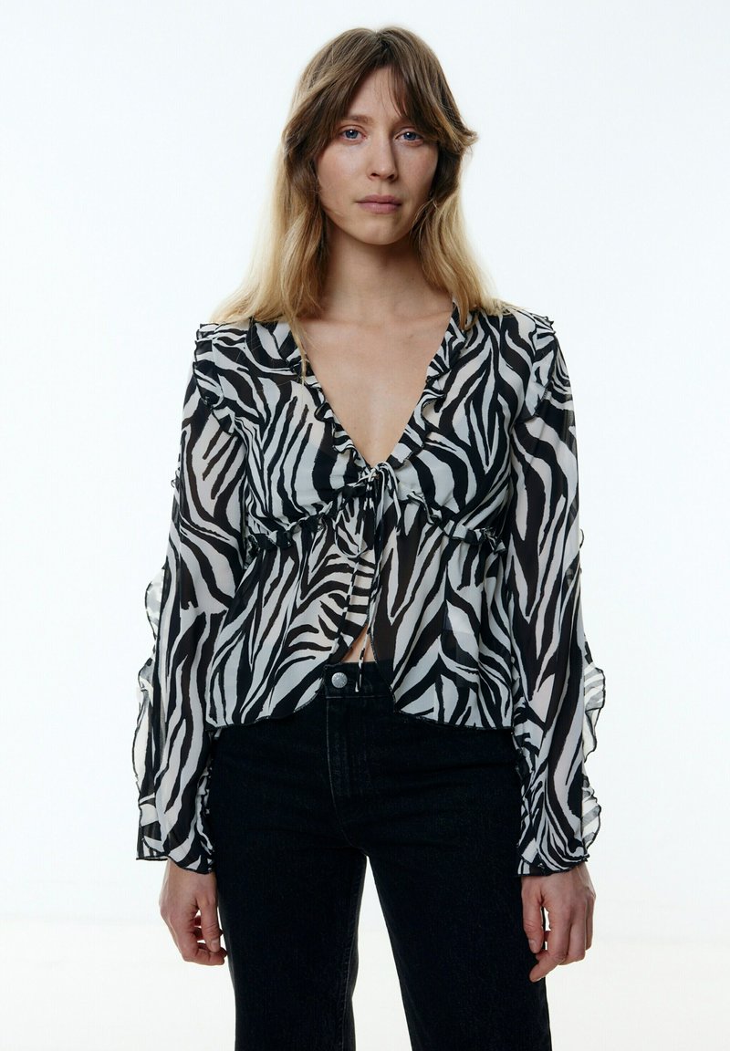 Durchscheinende Bluse im Schwarz-Weiß-Zebra-Druck mit gerafften Kanten, V-Ausschnitt und versammeltem Bindedetail an der Vorderseite, kombiniert mit schwarzen Jeans.