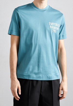 Man met een teal korte mouw t-shirt met de tekst "EMPORIO ARMANI 1981" op de borst en zwarte broek, staand tegen een effen achtergrond.