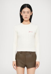 Biały sweter dzianinowy z wzorem w warkocze i prążkowanym wykończeniem, z czerwonym haftem "Fiorucci" na piersi. W połączeniu z brązowymi szortami.