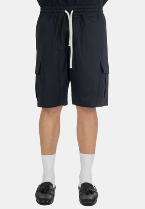 Persona che indossa pantaloncini cargo neri con cordino bianco, calzini bianchi a metà polpaccio e mocassini neri intrecciati con nappine.