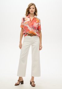 Chemise corail à manches courtes avec un motif floral, associée à un pantalon large blanc. Le mannequin porte des sandales à talons bruns et une ceinture assortie.