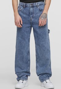 Light blue denim jeans with a loose fit, five pockets, and contrast stitching, paired with white sneakers. Textured fabric with a faded wash.