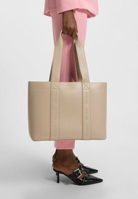 Beige kein nahka -tote -laukku, jossa on kohotettu "HUGO" -brändäys, jämäkkä muoto ja kaksoistasaista kahvat; yhdistettyinä vaaleanpunaisiin housuihin ja mustiin korkokenkiin.