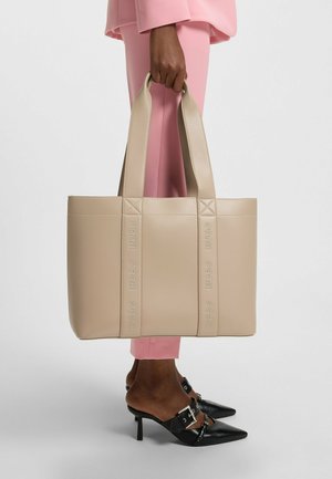 Beige faux leather tote bag with embossed "HUGO" branding, structured shape, and dual flat handles; paired with pink pants and black heels.