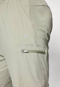 Pantalones cargo en un tono caqui claro con un bolsillo lateral con cremallera, cinturilla elástica y un logotipo bordado sutil. El material parece suave y duradero.