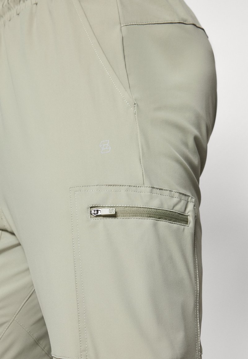 Pantaloni cargo color khaki chiaro con tasca laterale con zip, fascia elastica in vita e logo ricamato sottile. Il materiale sembra liscio e resistente.