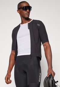 Veste de cyclisme noire de performance et short noir ajusté, associés à un t-shirt en mesh blanc. Les accessoires comprennent un casque noir et des lunettes de soleil sombres.