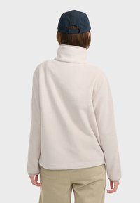 Sudadera de forro polar blanca con cuello alto, textura suave y corte relajado. Combinada con pantalones de color claro y una gorra azul marino.