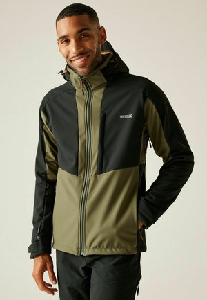 Uśmiechnięty mężczyzna ubrany w czarną i oliwkowozieloną kurtkę outdoorową na suwak z kapturem, stojący z jedną ręką w kieszeni na neutralnym tle.