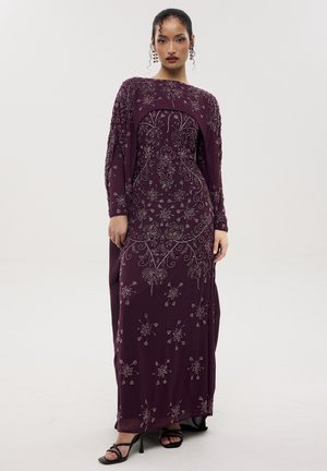 Femme portant une longue robe violette foncée avec une broderie florale complexe en perles et des boucles d'oreilles assorties, debout devant un fond clair uni.