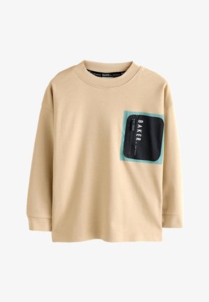 Garçon debout et souriant, portant une chemise beige à manches longues avec une poche zippée noire sur la poitrine et un pantalon foncé.