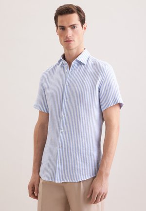 Homme portant une chemise à manches courtes à rayures verticales bleu clair et blanc avec un pantalon beige, debout devant un fond uni.