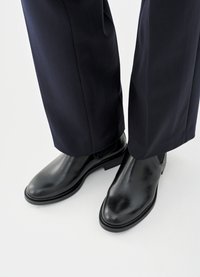 Bottes Chelsea en cuir noir avec une finition lisse, semelles plates et panneaux latéraux élastiques, associées à un pantalon sombre sur mesure.