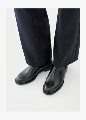 Zwarte leren Chelsea-enkellaarzen met een glanzende afwerking, platte zolen en elastische zijkanten, gecombineerd met donker, op maat gemaakte pantalons.