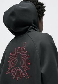 Μαύρο φούτερ με κου κούλα που διαθέτει μεγάλο κόκκινο κεντημένο λογότυπο Jumpman και κυκλικό σχέδιο στην πλάτη. Απαλή ύφανση με κανονική εφαρμογή και κουκούλα.