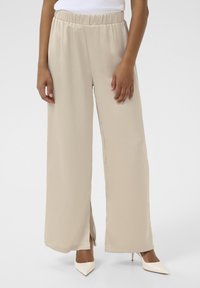 Pantalons beige à jambes larges avec une taille élastique, texture lisse et fentes sur les côtés, associés à des talons aiguilles blancs.