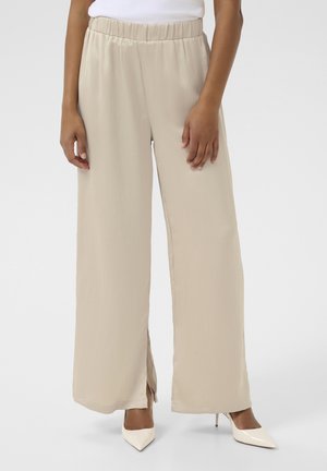 Pantalons beige à jambes larges avec une taille élastique, texture lisse et fentes sur les côtés, associés à des talons aiguilles blancs.