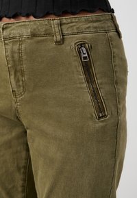Olive green pants made of soft fabric, featuring a zippered side pocket, standard pockets, and subtle textured detailing.