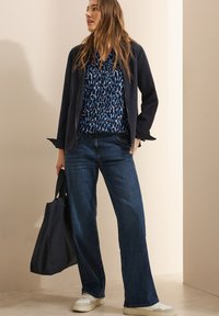 Giacca blu navy, blusa stampata blu e jeans a gamba larga in denim scuro; modella tiene una borsa a tracolla blu navy; sneakers bianche completano il look.