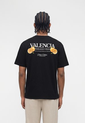 Fonott hajú férfi háttal, fekete pólót visel, amelyen a "VALENCIA NARANJO VIEJO" felirat és narancs gyümölcs grafikák láthatók a háton.