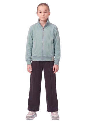 Une fille se tient devant un fond blanc, vêtue d’une veste vert pâle zippée, d’un pantalon foncé et de baskets roses et grises.