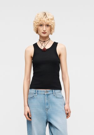 Person med krøllet blondt hår iført en sort ærmeløs top, blå jeans og et sort halskæde med en rød rosevedhæng, står mod en grå baggrund.