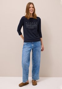 Navyblaues Langarmshirt mit grafischem Text, kombiniert mit hellblauen weitgeschnittenen Jeans und braunen Slippern, vor einem neutralen Hintergrund.
