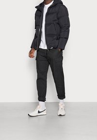 Mężczyzna ubrany w czarną kurtkę puchową, białą koszulkę, ciemne spodnie cargo podwinięte przy kostkach, białe skarpetki oraz białe sneakersy Nike z czarnym logo.