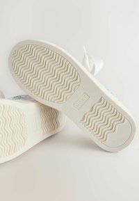 Semelle en caoutchouc blanche avec un motif texturé en vagues ; présente la marque "NEXT" près du talon. Design fin et plat adapté à une tenue décontractée.