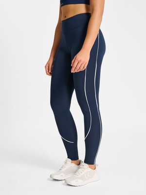 Frau trägt marineblaue Leggings mit hohem Bund mit weißen geschwungenen Linien und weißen Sportschuhen, steht vor einem einfarbigen Hintergrund.