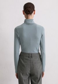 Frau mit dunklem Haar im Dutt, die einen hellblauen Rollkragenpullover und graue hochgeschnittene Hose trägt, steht vor einer weißen Wand.