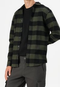 Grøn og sort ternet flannel-skjorte med knapper foran og krave. Kombineret med en sort t-shirt og grå cargobukser. Tekstureret stof.