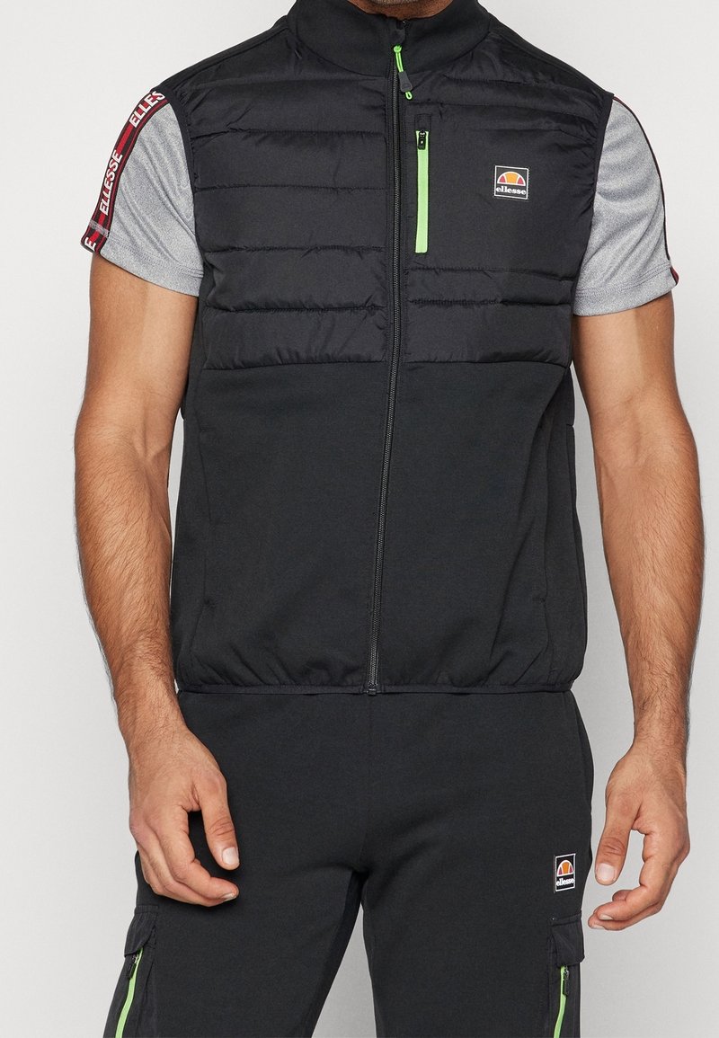 Czarna pikowana kamizelka z wysokim kołnierzem, zamkiem błyskawicznym z przodu i zielonymi detalami zamka. Pod spodem szara koszula z krótkim rękawem z logo na rękawach.