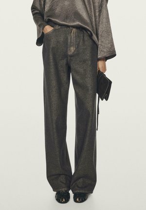 Person wearing loose dark gray pants and a shiny long-sleeve top, holding a small black clutch in one hand, with black open-toe shoes.