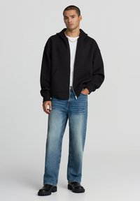 Felpa nera con zip realizzata in tessuto morbido, indossata sopra una camicia bianca, abbinata a jeans a gamba dritta azzurri chiari e scarpe chunky nere.