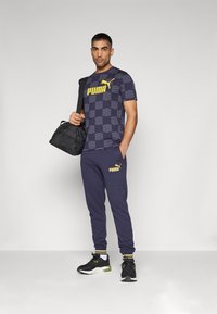 Camiseta deportiva de rayas marinas con logo amarillo, combinada con pantalones de chándal marinos que presentan el logo y zapatillas deportivas negras. Mochila negra al hombro.