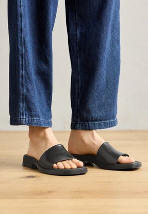 Sandalias deslizantes de cuero negro con una tira superior ancha y punta abierta; combinadas con pantalones de mezclilla azules, situadas sobre un suelo de madera.