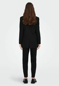 Svart kavaj med pinstripes och strukturerade axlar, ihop med åtsittande svarta byxor och kraftiga svarta skor. Långt hår faller nerför ryggen.